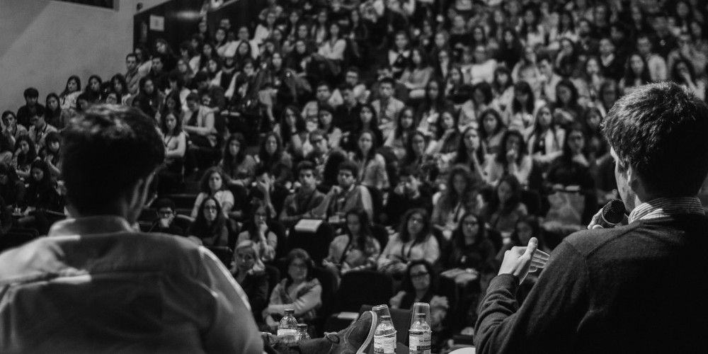 View from behind two speakers sat on stage. One speaking into a microphone before a large crowd in a lecture hall.